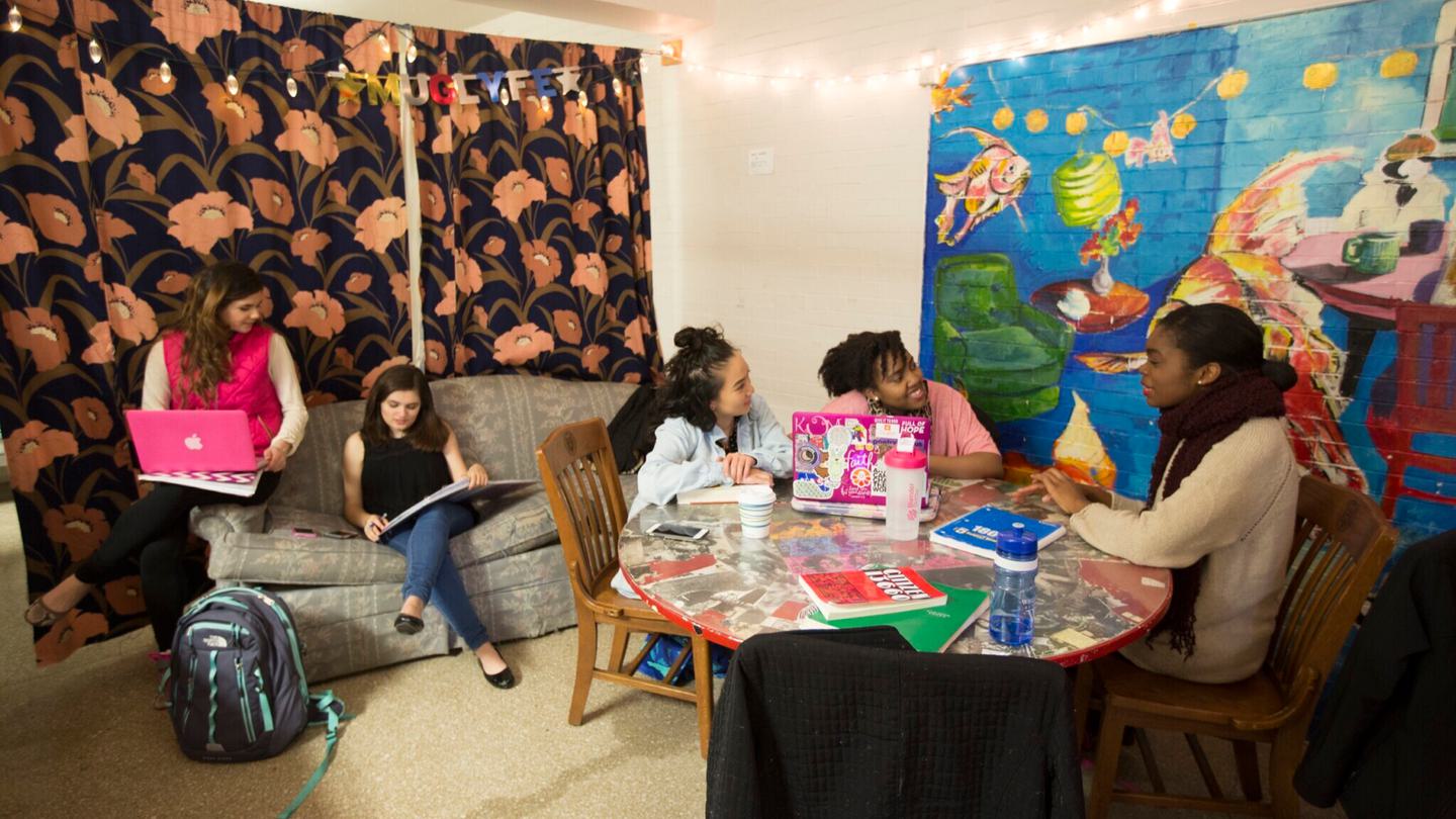 Two students sit a couch. Three students sit at a table. The walls are covered with a colorful mural and flower curtain.