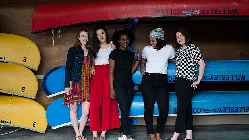 Five Wellesley College students pose for a photo in front of blue canoes in a boat house