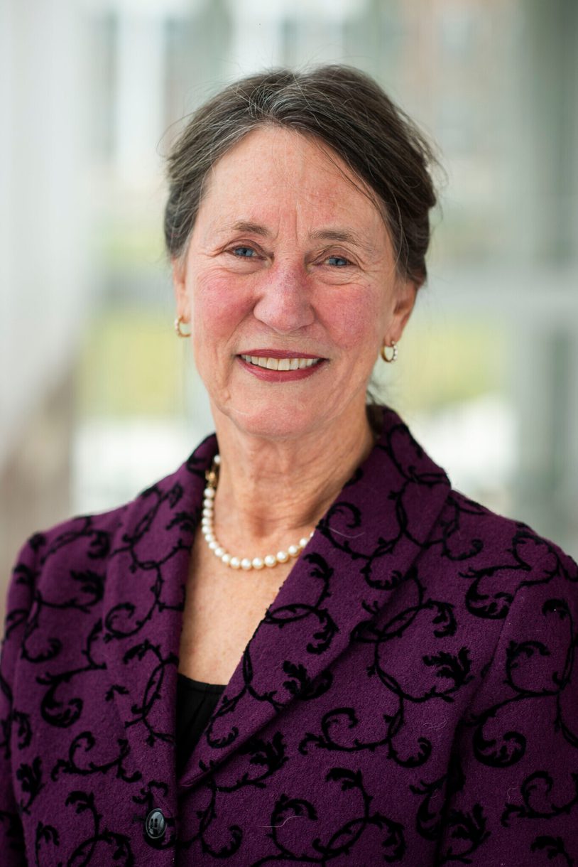 A head shot of a woman with her hair pulled back, wearing a pearl necklace and a plum-colored jacket.