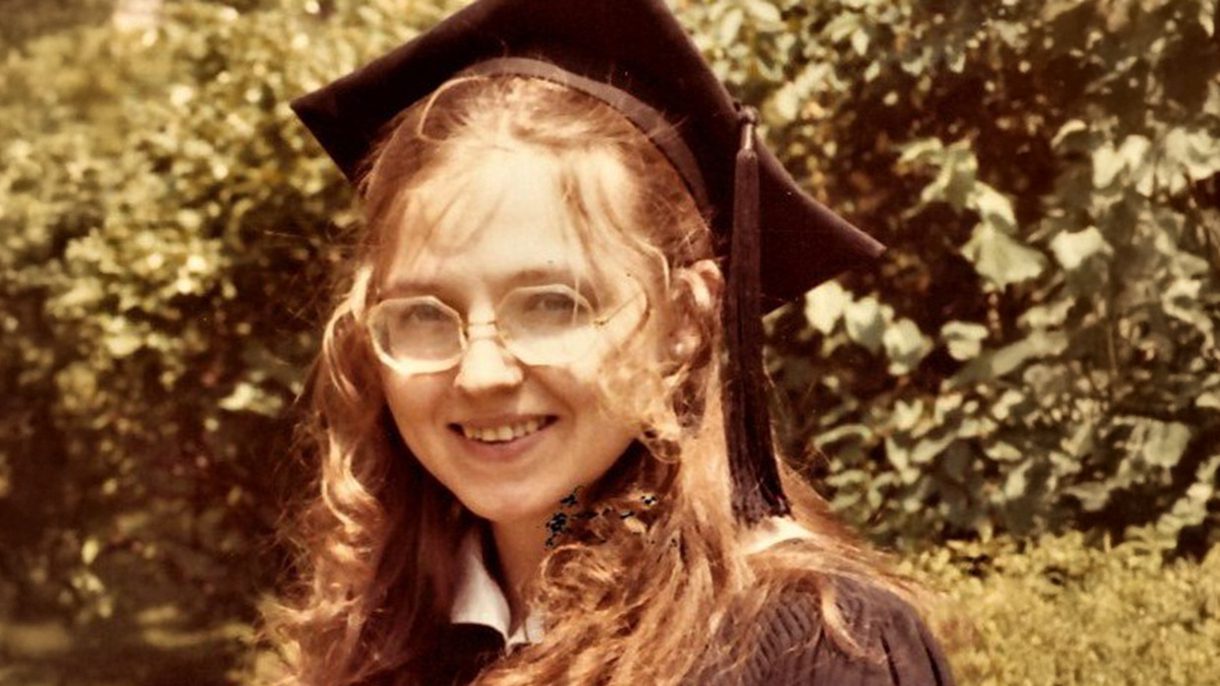 A young woman with long hair and wearing glasses and a graduation mortarboard smiles at the camera.