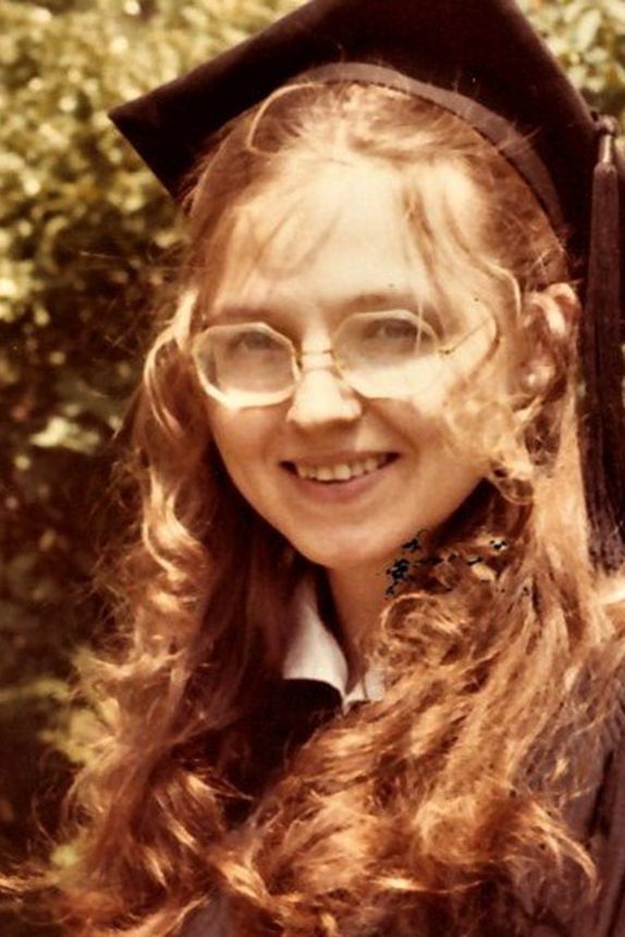 A young woman with long hair and wearing glasses and a graduation mortarboard smiles at the camera.