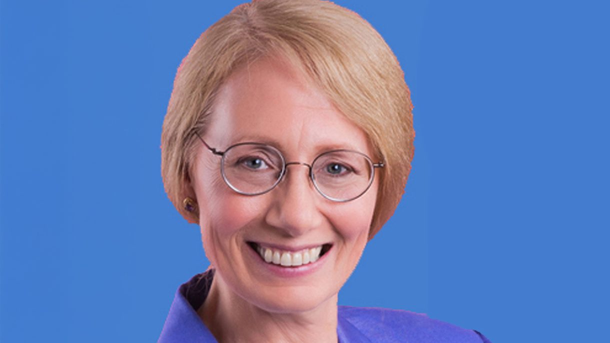 Woman with short blonde hair and glasses smiles at the camera. She is in front of a blue background.