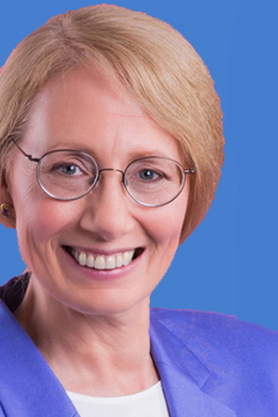 Woman with short blonde hair and glasses smiles at the camera. She is in front of a blue background.