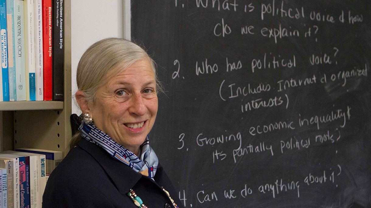 A woman smiles at the camera while standing near a classroom blackboard.