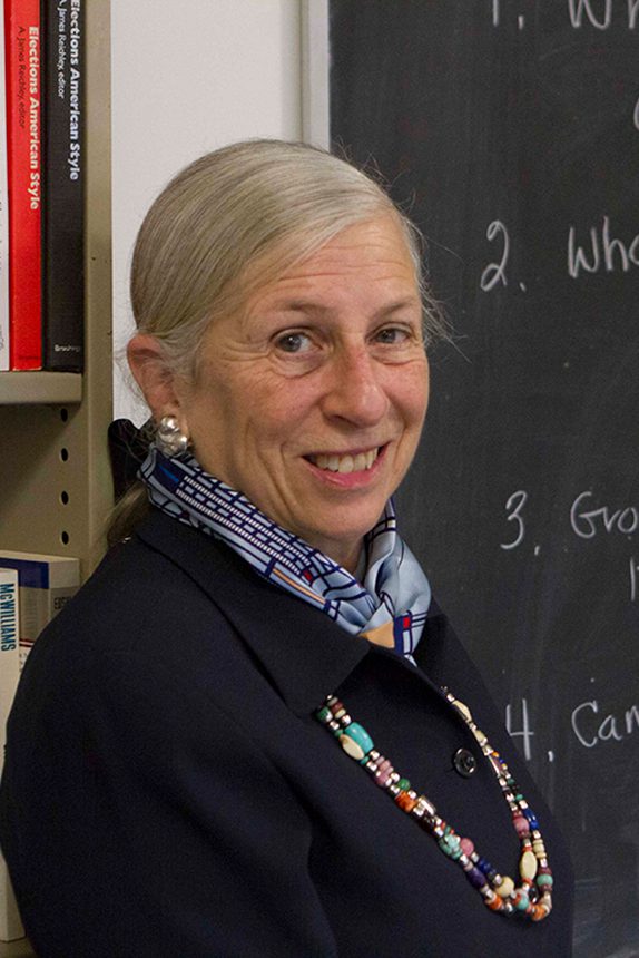 A woman smiles at the camera while standing near a classroom blackboard.