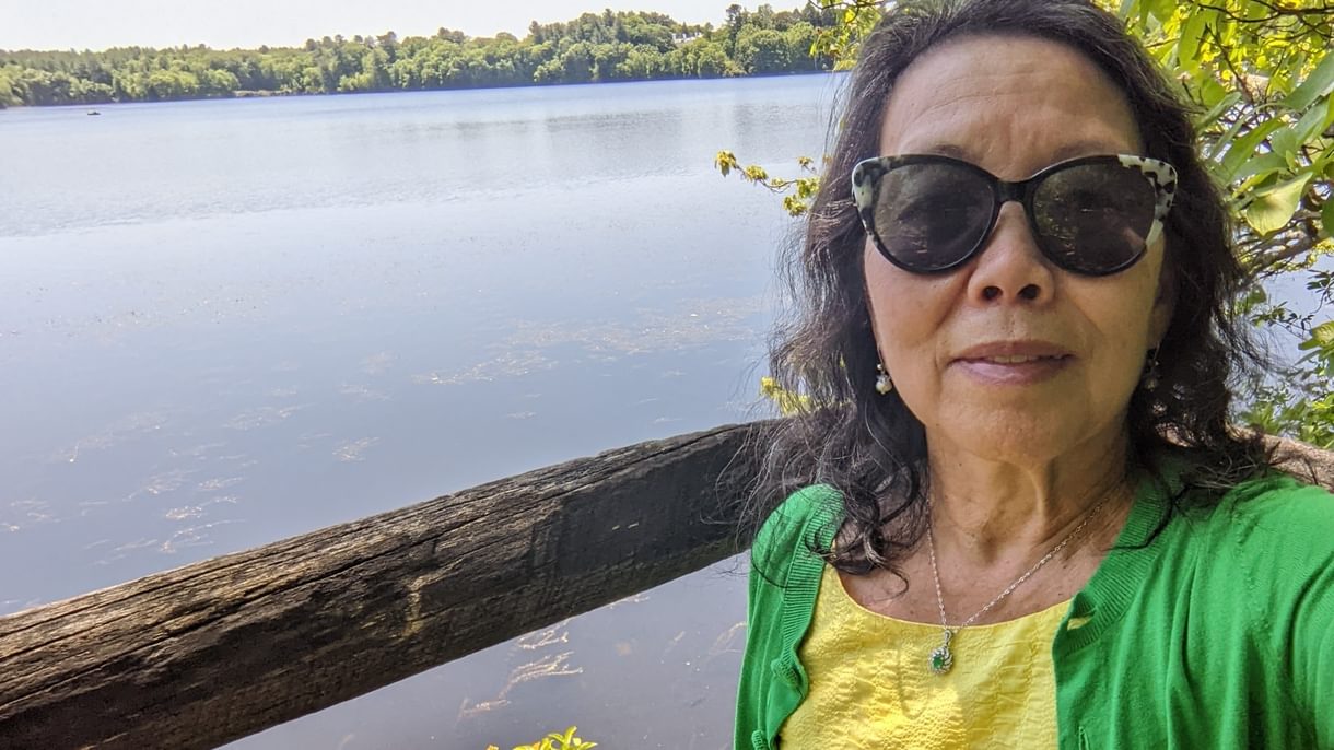 A woman in a yellow dress with a green sweater takes a selfie while sitting on a bench near Lake Waban.