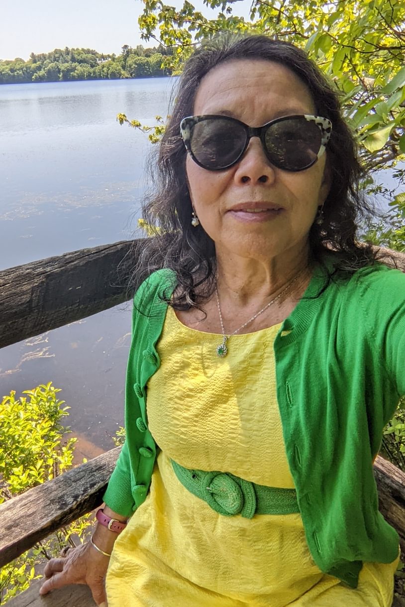 A woman in a yellow dress with a green sweater takes a selfie while sitting on a bench near Lake Waban.