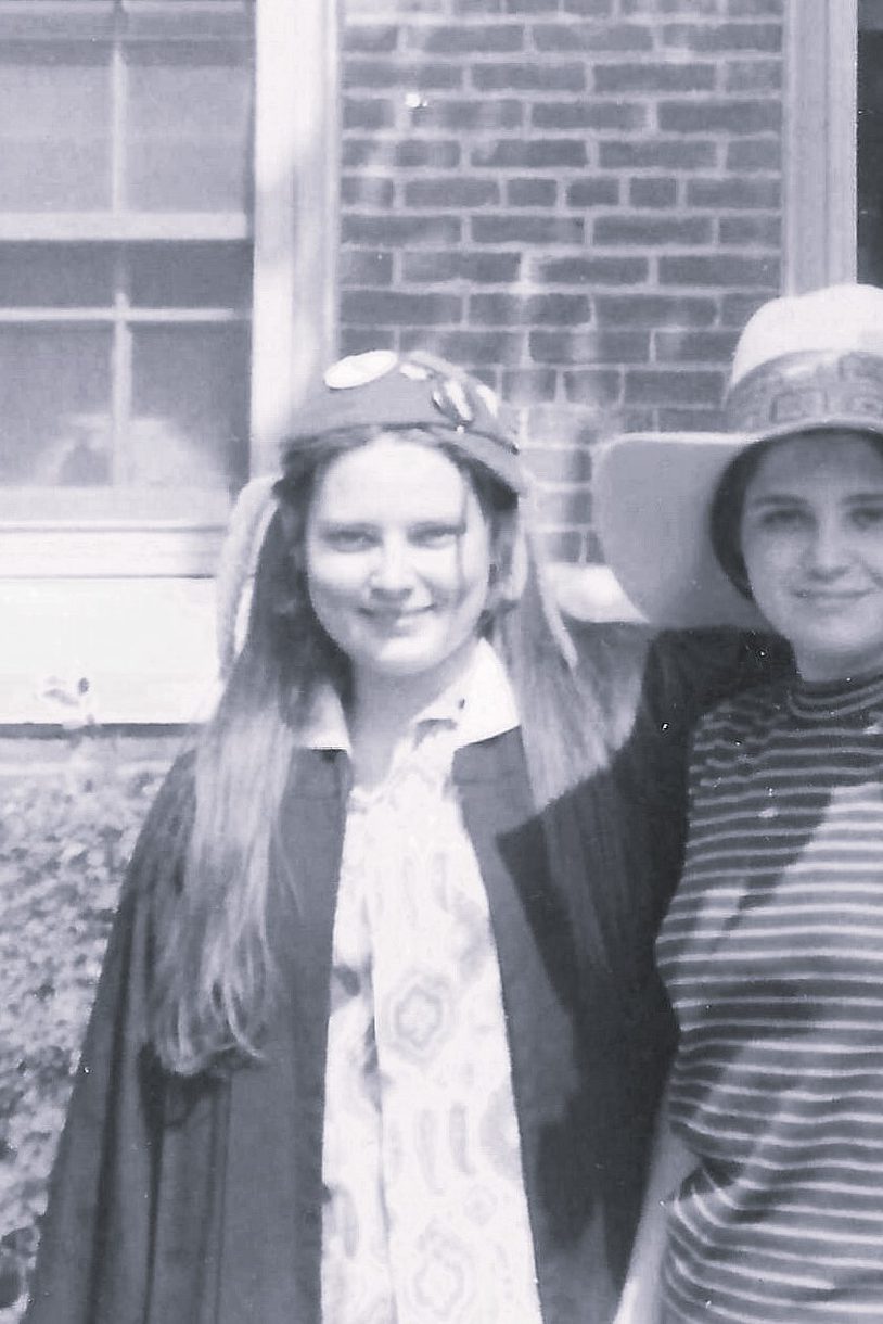Black and white image of two young women standing, looking at camera. One has her arm on the shoulders of the other. Both wear hats.