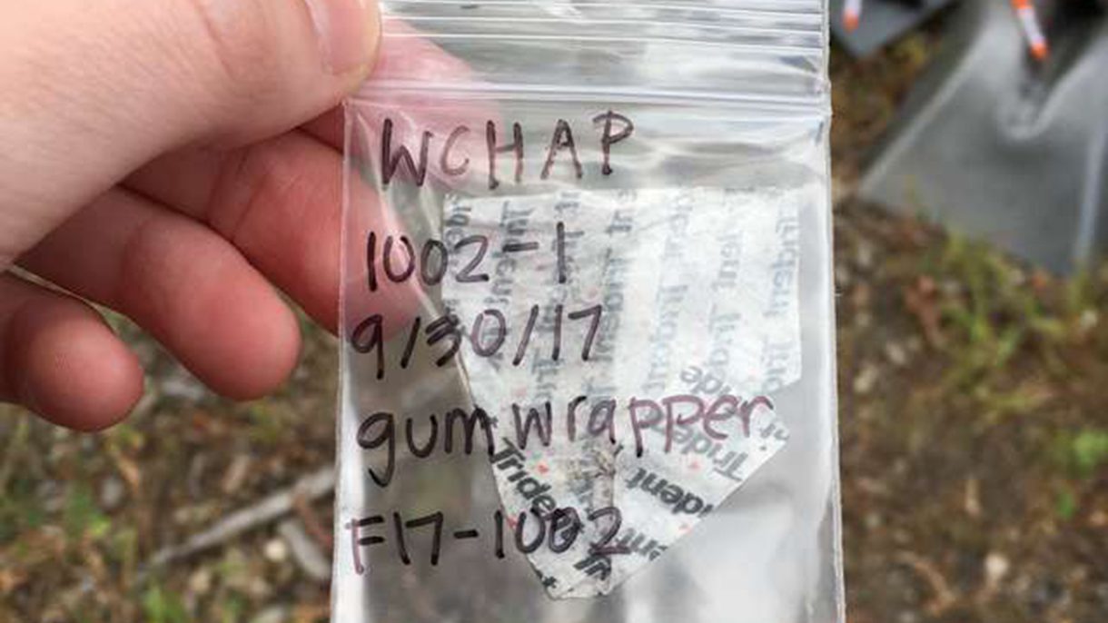 A hand holds a small plastic bag with a gum wrapper inside. On the bag is written WCHAP 1002-1 9/30/2017 gum wrapper f17-1002