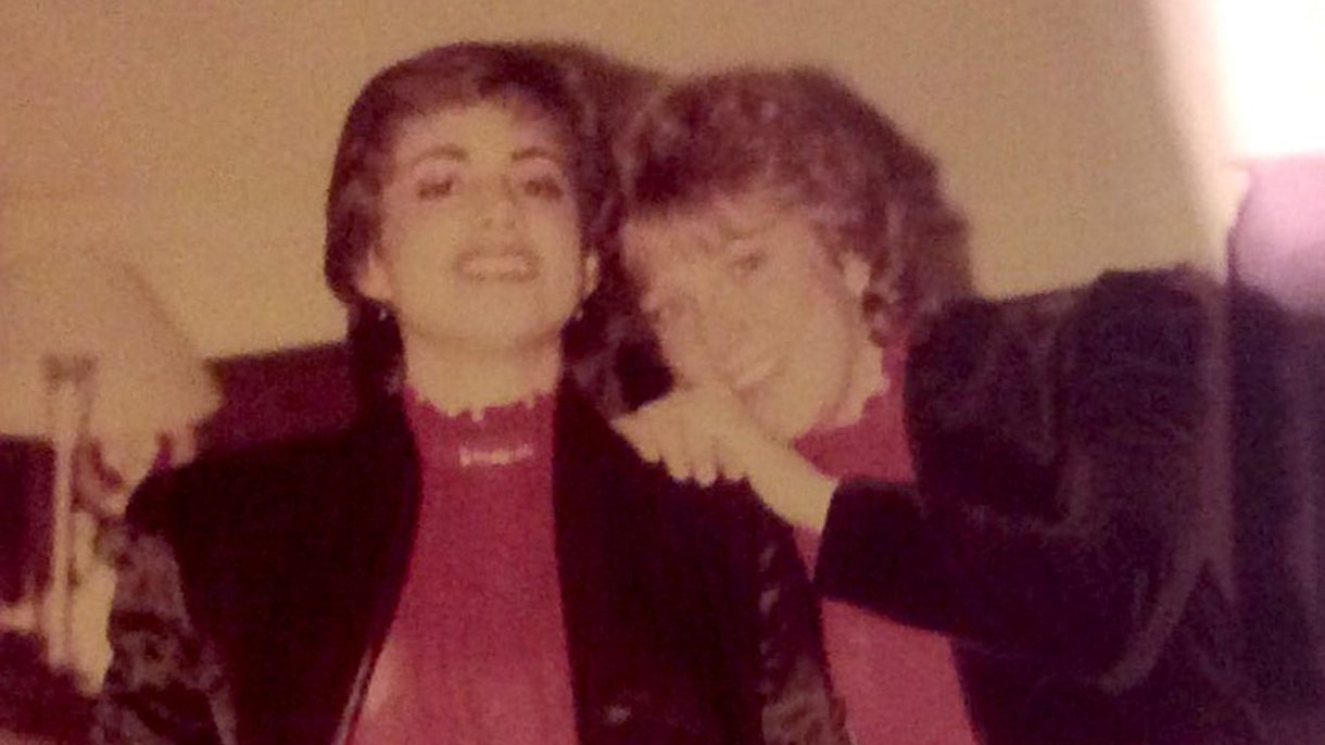 Two women wearing matching jackets and tops pose for a picture, with one holding the shoulders of the other and leaning in.