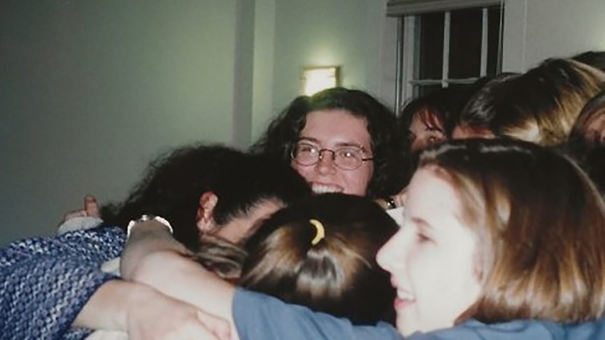 A group of young women cluster together in ball with their arms around each other. One face is visible in the center, looking at the camera. One face is visible in profile, looking to the left.