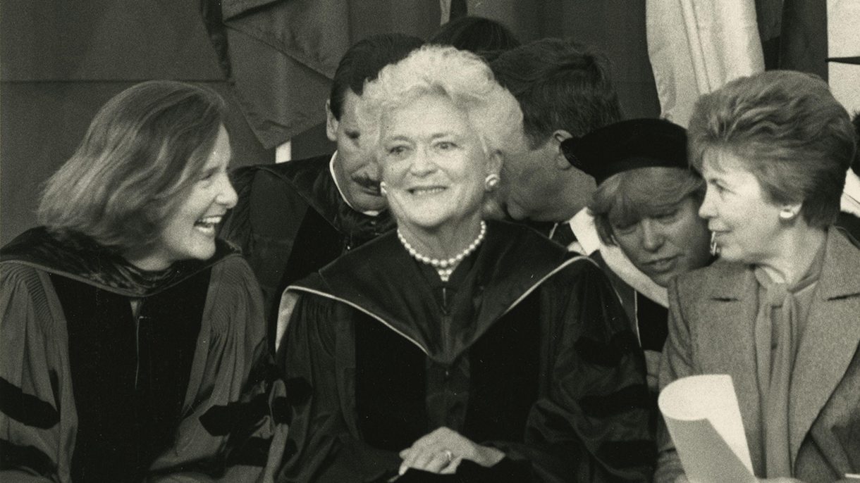 Barbara Bush sits smiling in regalia and flanked by two women smiling at each other.