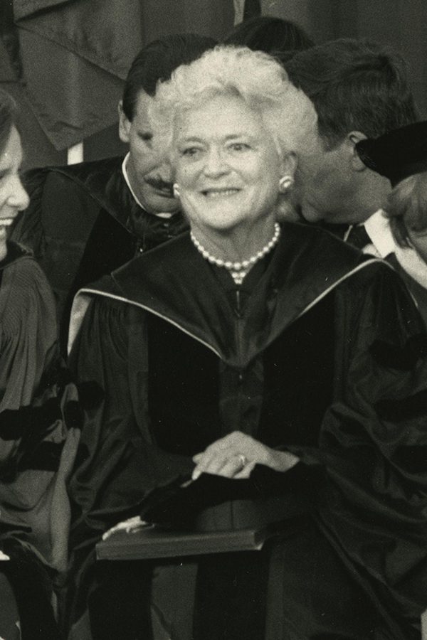 Barbara Bush sits smiling in regalia and flanked by two women smiling at each other.
