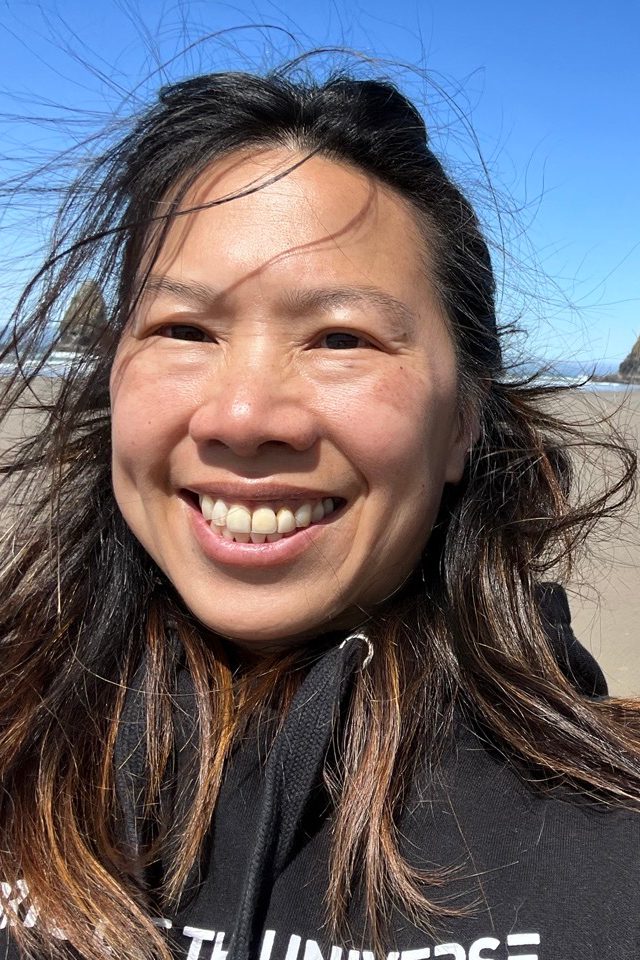 A woman with long dark hair takes a selfie on a coastline with a large rock in the background.