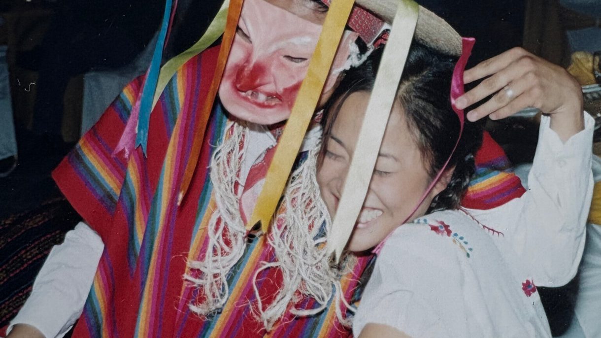 A womans laughs as a person in a poncho and sombrero and mask sits on her lap.