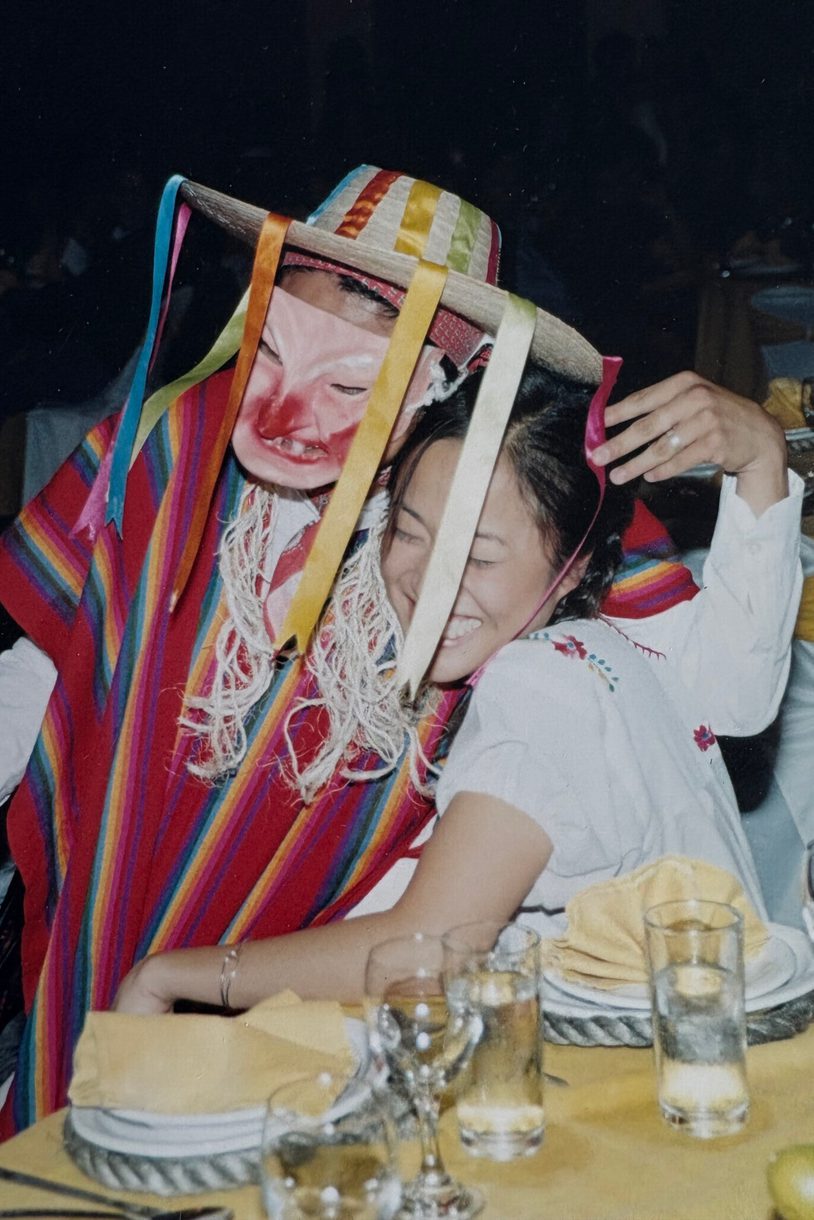 A womans laughs as a person in a poncho and sombrero and mask sits on her lap.