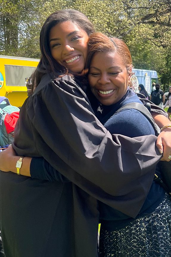 A student wearing a graduation gown hugs her professor.