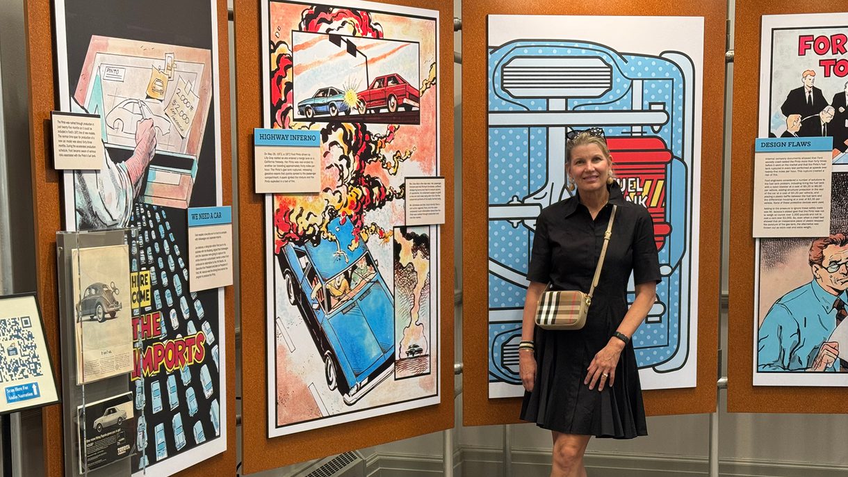 A woman in a black dress stands in front of an exhibition showing comic-style art work.