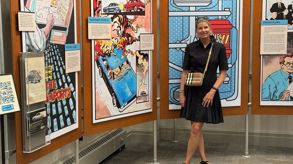 A woman in a black dress stands in front of an exhibition showing comic-style art work.