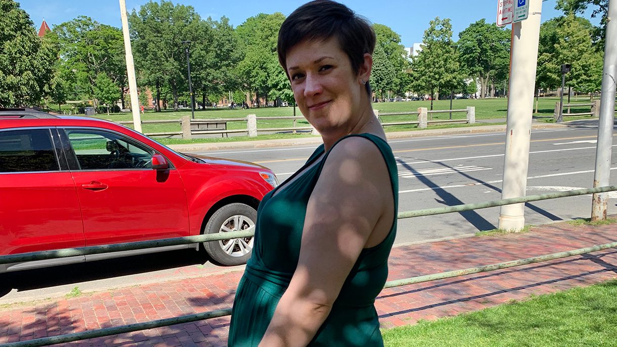 A woman in a green dress stands in profile with her hands in front of her dress showing her pregnant belly.