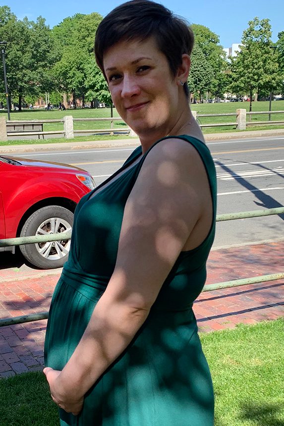 A woman in a green dress stands in profile with her hands in front of her dress showing her pregnant belly.