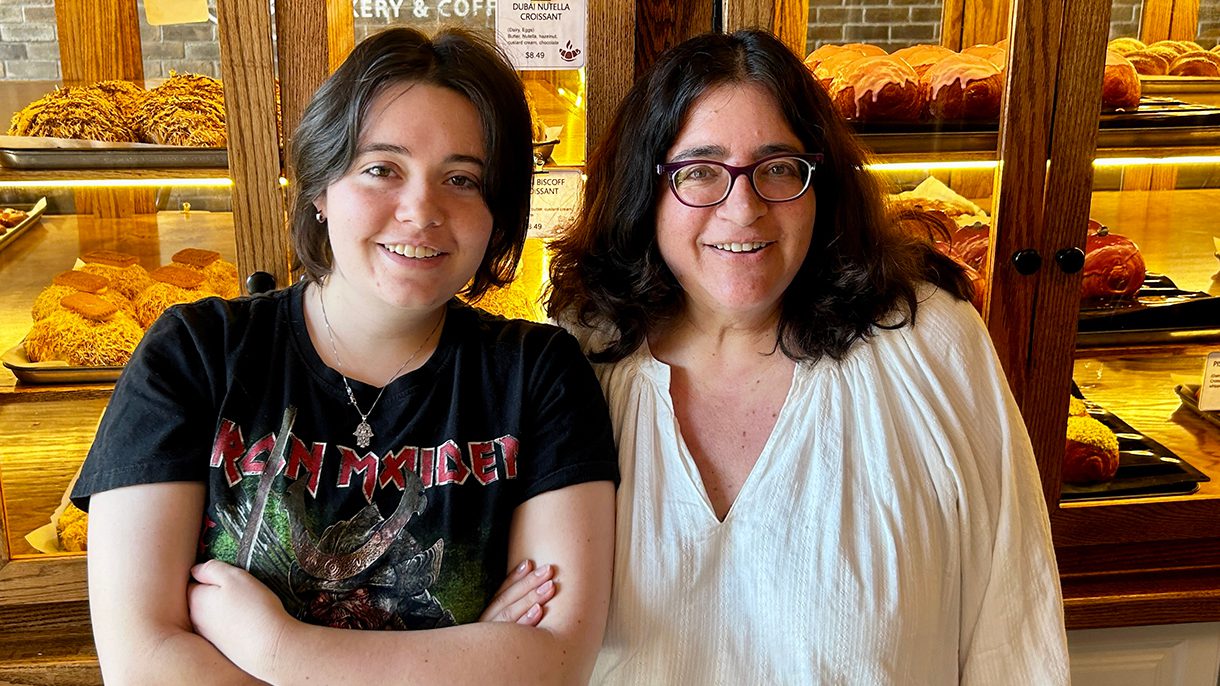 A mom and her daughter stand in front of a case of baked goods, smiling at the camera.