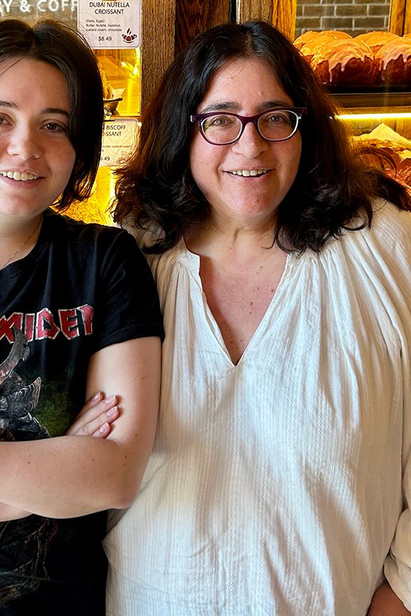 A mom and her daughter stand in front of a case of baked goods, smiling at the camera.