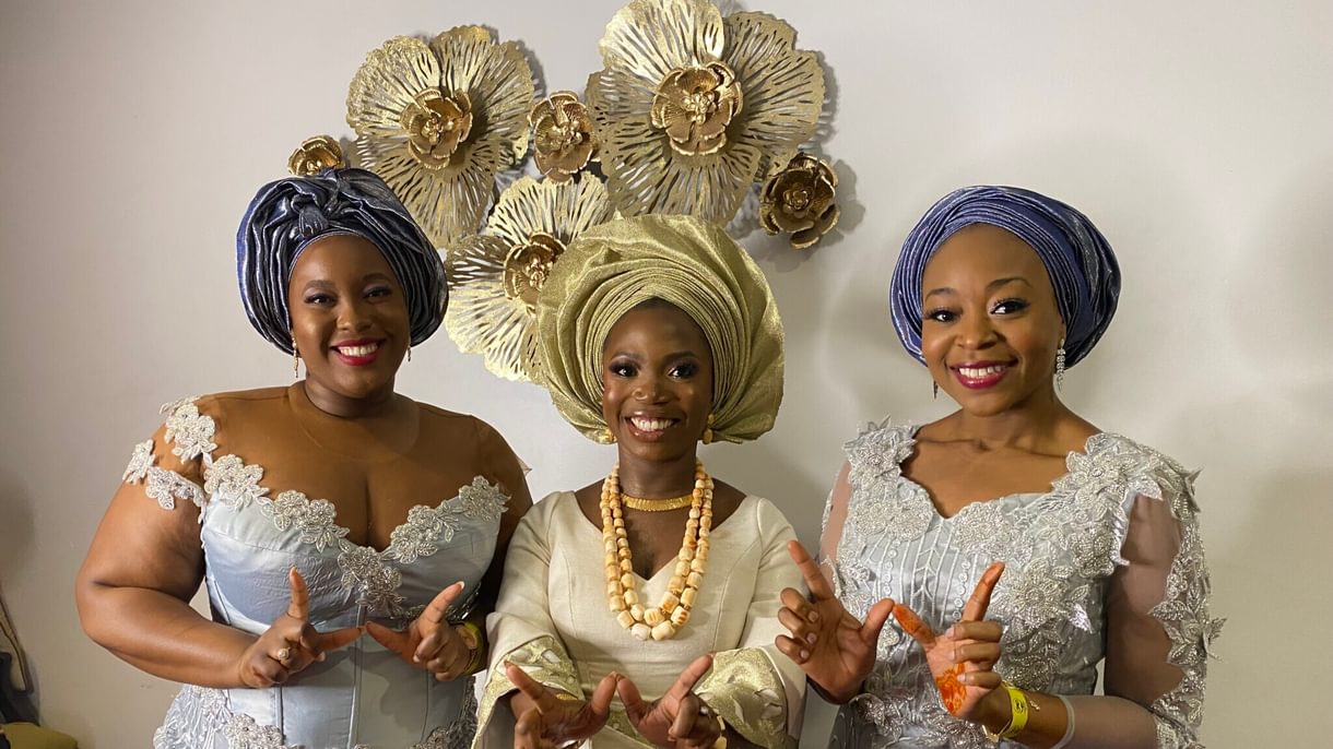 Three women wearing formal dresses and head turbans each hold their hands up to form a W.