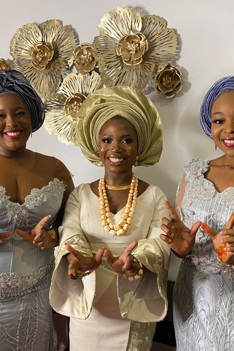 Three women wearing formal dresses and head turbans each hold their hands up to form a W.