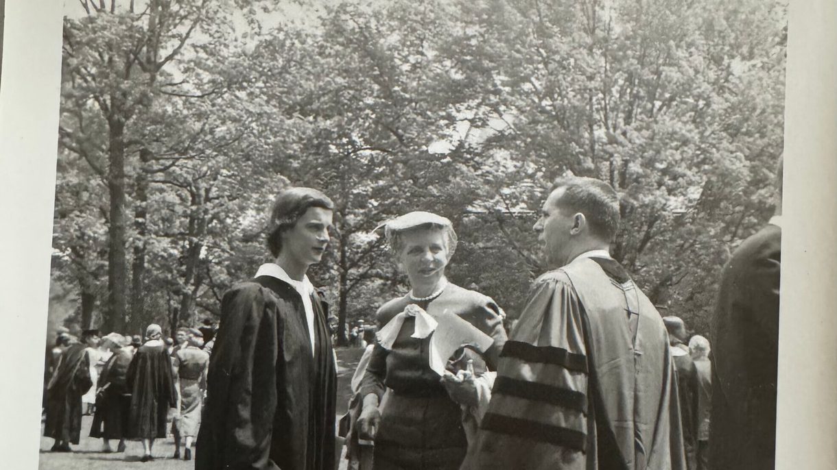 A black and white photo showing a woman in graduate regalia facing a man in regalia. In between them is a woman wearing a hat with a veil over her face and a dark dress.