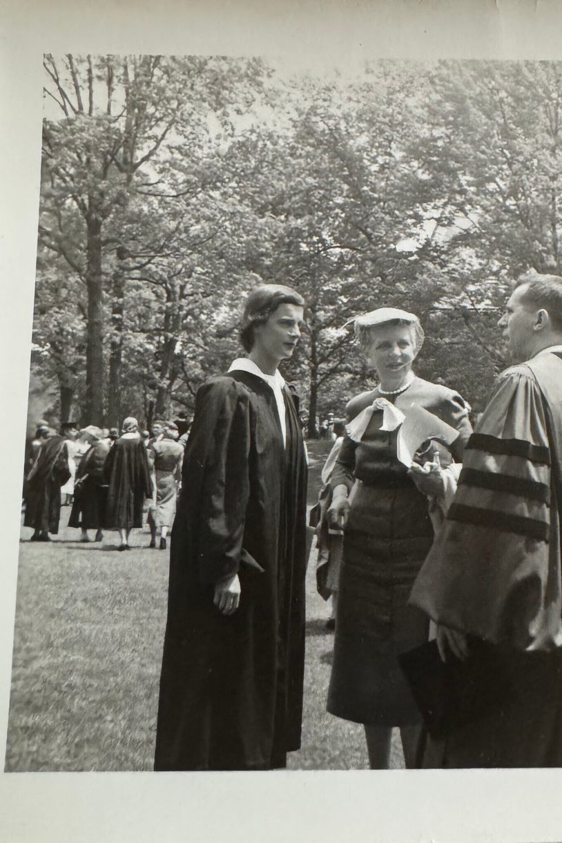 A black and white photo showing a woman in graduate regalia facing a man in regalia. In between them is a woman wearing a hat with a veil over her face and a dark dress.