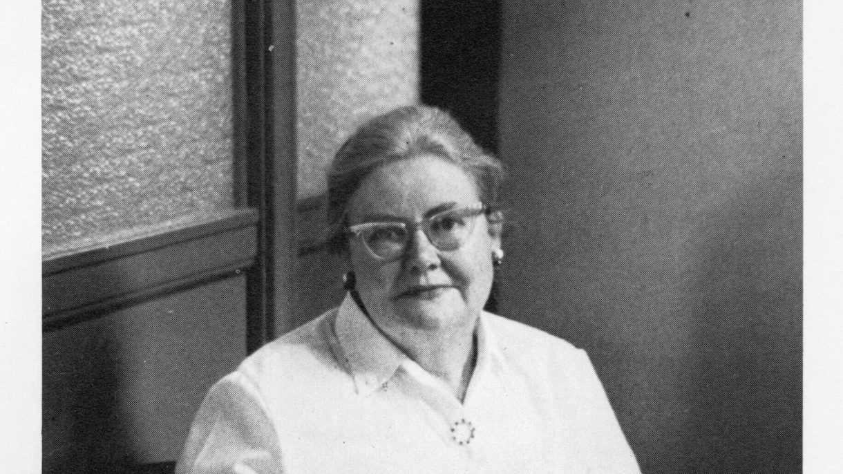 A black and white photo of a woman seated, looking at the camera and holding a book in her lap.