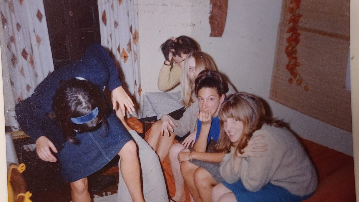 One woman stands with her foot on the belly of another woman lying on the floor. Four women sit on a couch looking serious and some of them look at the camera