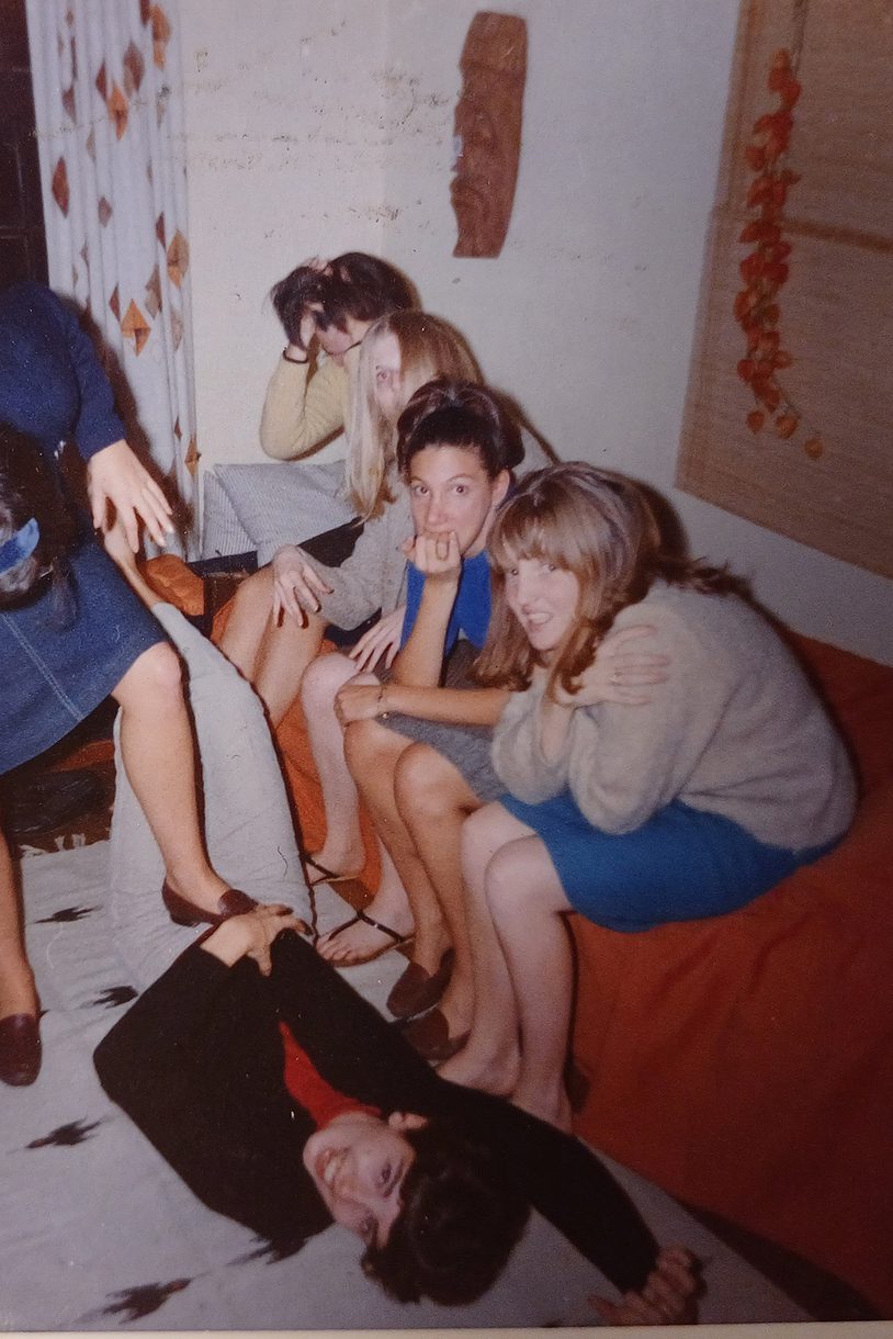 One woman stands with her foot on the belly of another woman lying on the floor. Four women sit on a couch looking serious and some of them look at the camera