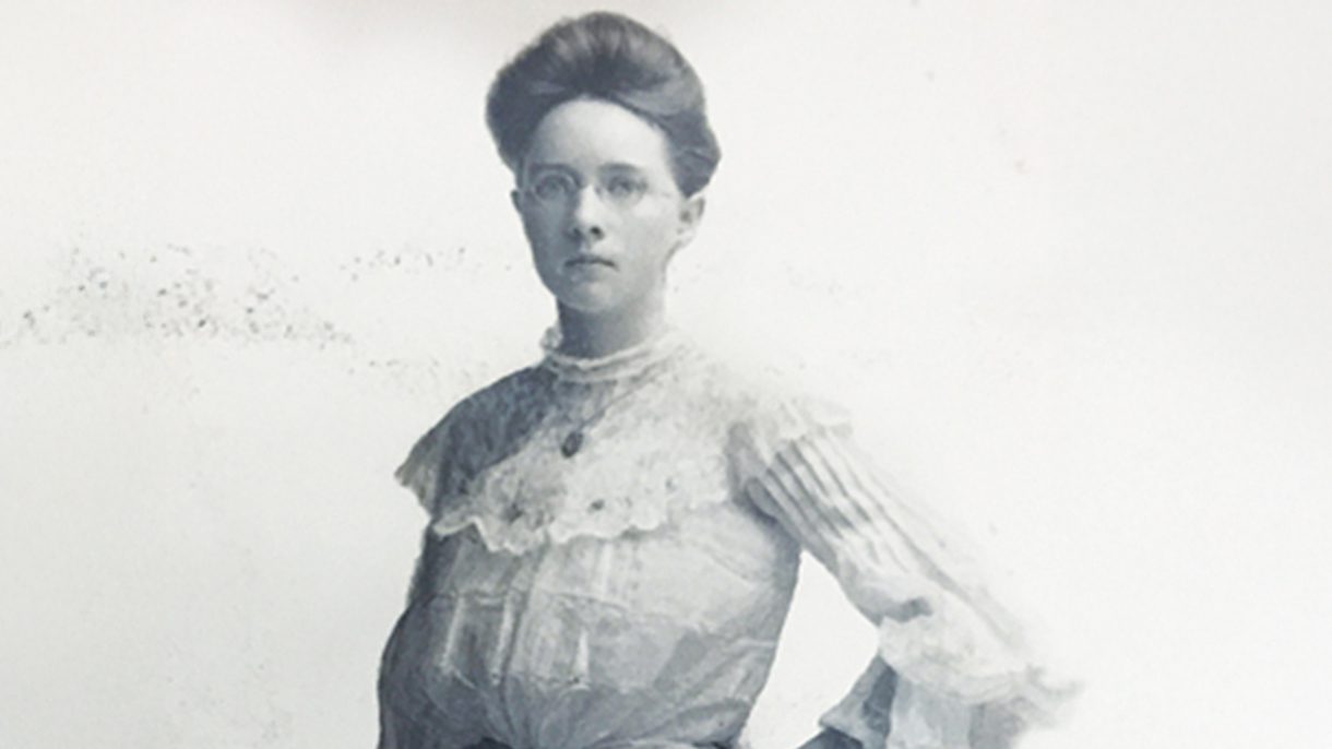 An archive photos of a woman standing straight, hand on her hip and holding a rolled up piece of paper. Her hair is pulled back.
