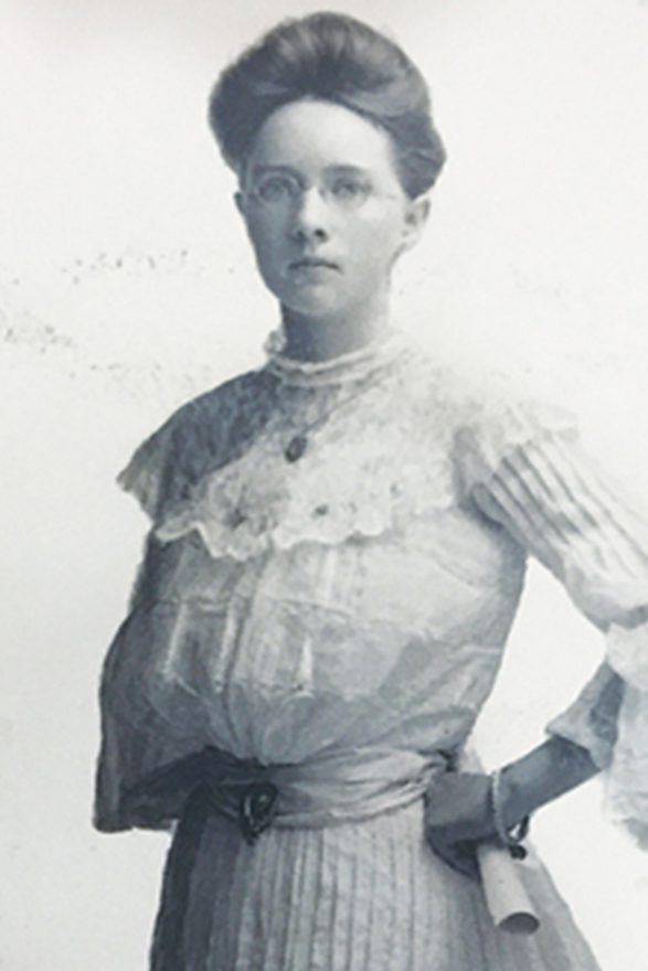 An archive photos of a woman standing straight, hand on her hip and holding a rolled up piece of paper. Her hair is pulled back.