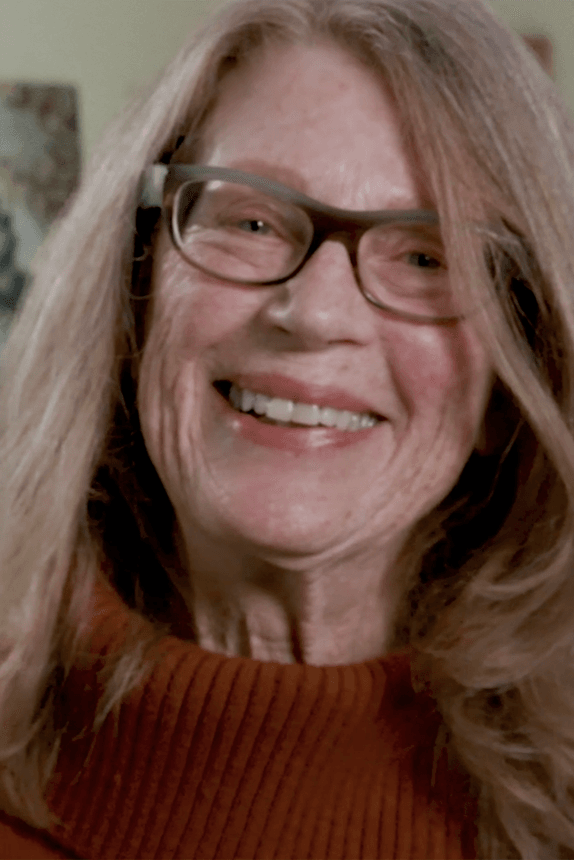 A woman looks at the camera and smiles. She has shoulder-length hair that frames her face and wears glasses.