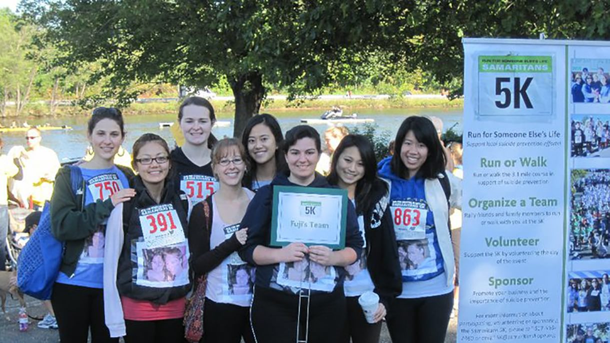 A group of women stand together next to a 5K sign with details about the race. They each have a racing bib on and a photo of two people taped to their shirts.