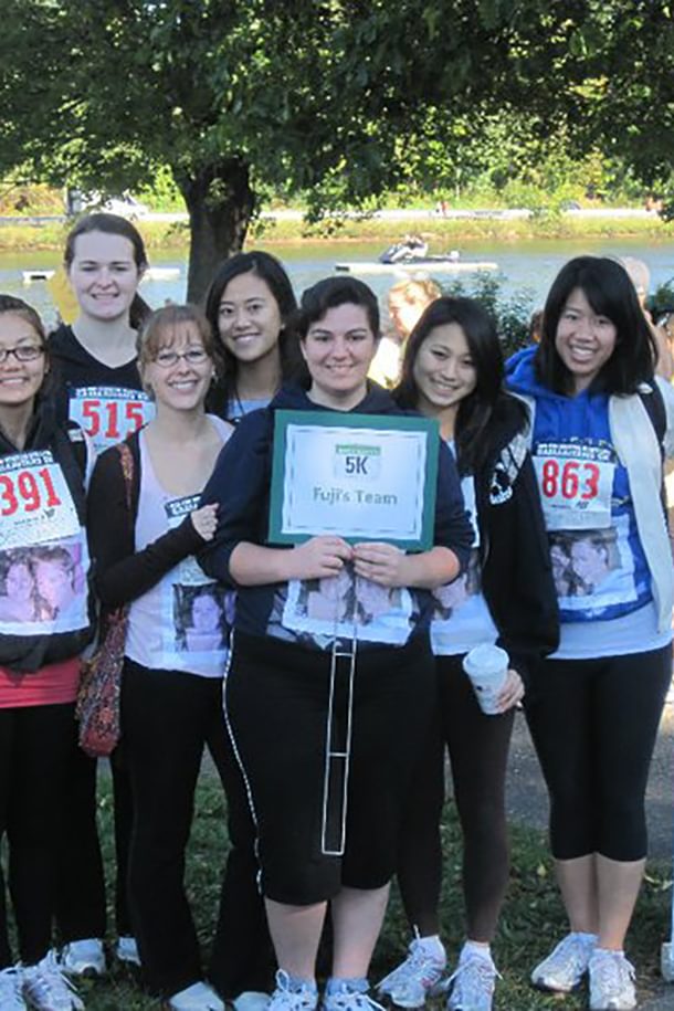 A group of women stand together next to a 5K sign with details about the race. They each have a racing bib on and a photo of two people taped to their shirts.