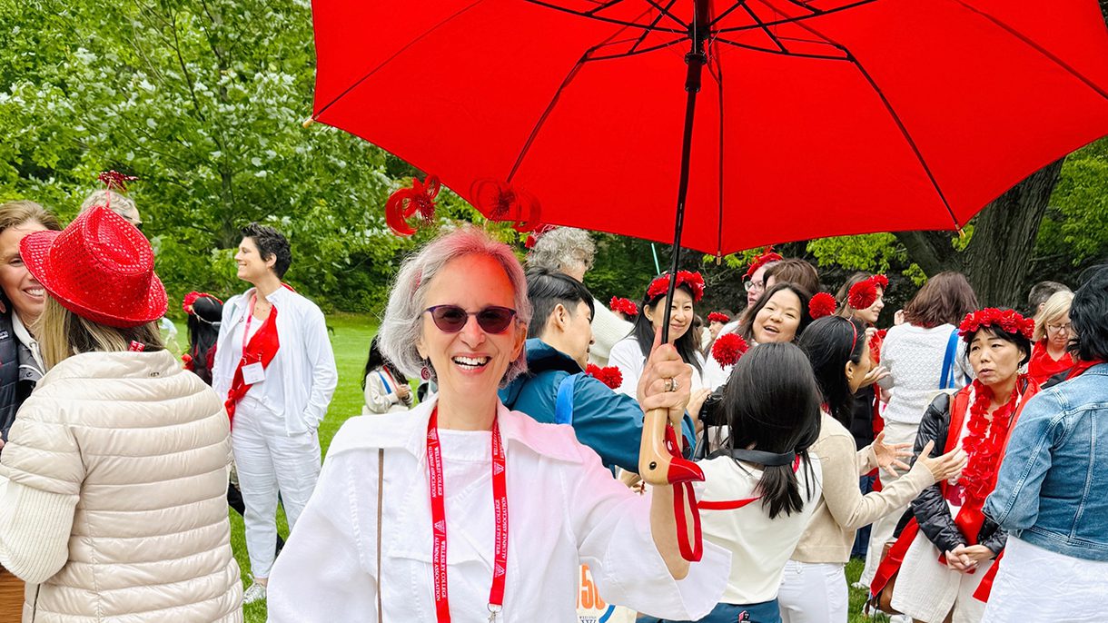 A woman dressed in white and holds a red umbrella. Behind her are others dressed in white with red hats or flowers.