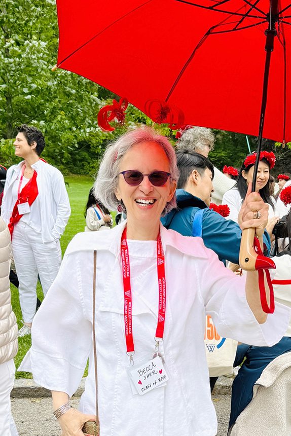 A woman dressed in white and holds a red umbrella. Behind her are others dressed in white with red hats or flowers.