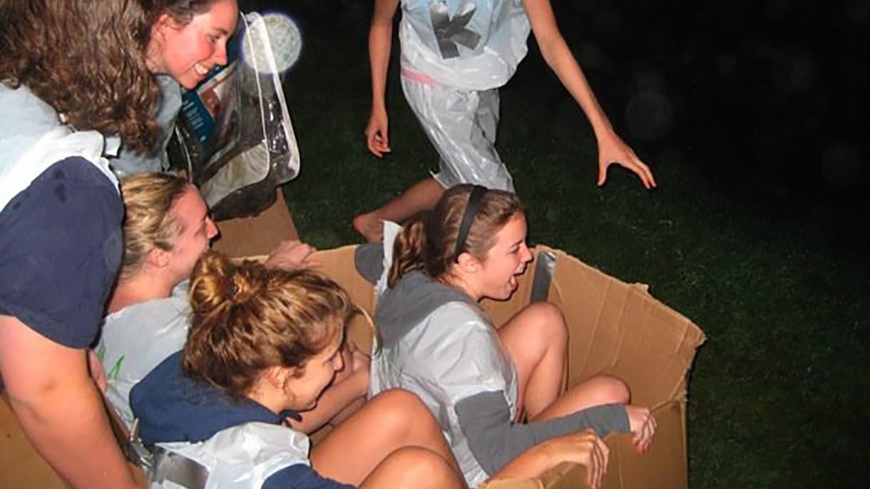 An action shot of two women inside a cardboard box, surrounded by classmates who are preparing to push them down a hill.