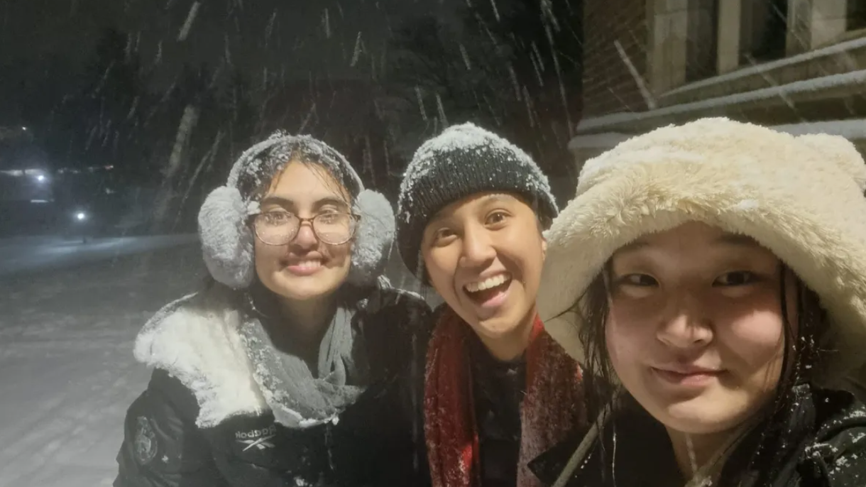 Three students wear winter clothes and smile at the camera. Snow falls around them.