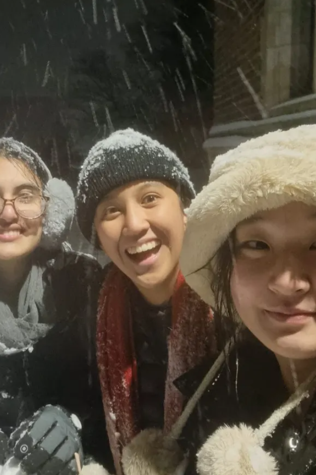 Three students wear winter clothes and smile at the camera. Snow falls around them.