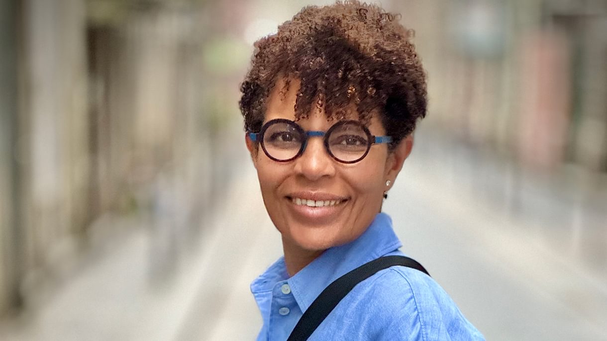A portrait of a woman in a blue button-up shirt, turned to the side and looking toward the camera. She wears glasses, short, curly hair, and a purse slung across her shoulder.