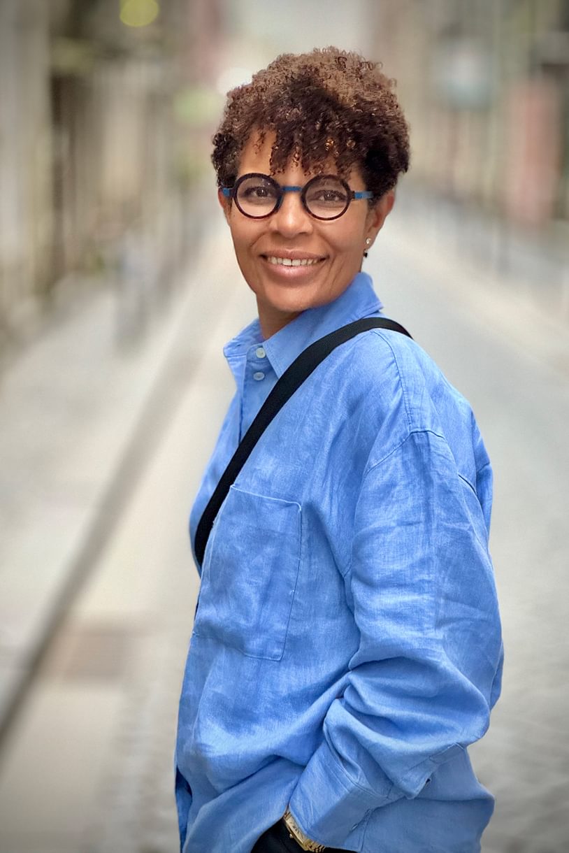 A portrait of a woman in a blue button-up shirt, turned to the side and looking toward the camera. She wears glasses, short, curly hair, and a purse slung across her shoulder.