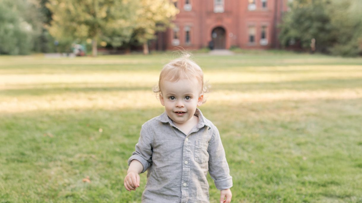 A toddler stands on a the Wellesley campus with a red brick campus building in the background.