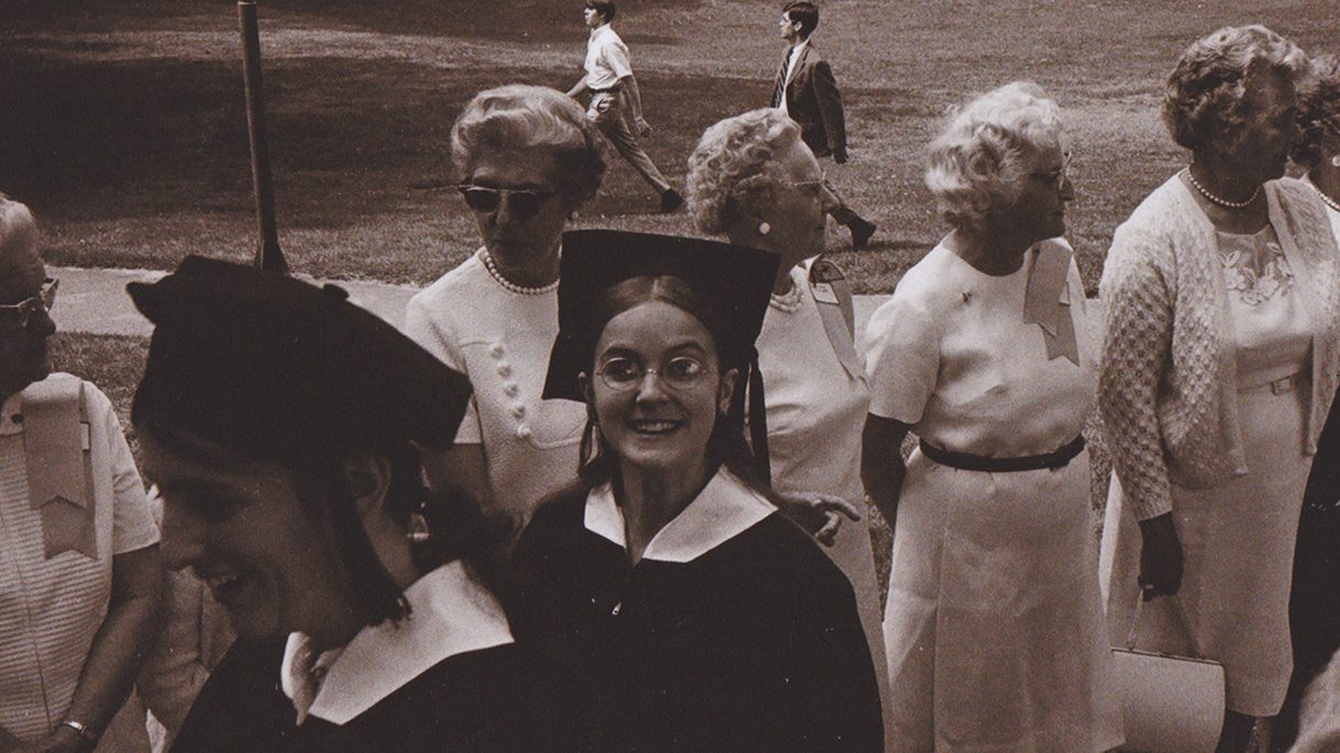 A young woman wearing her commencement gown and mortar board looks up at the camera as she processes during graduation. Behind her is a line of alumnae dressed in white.