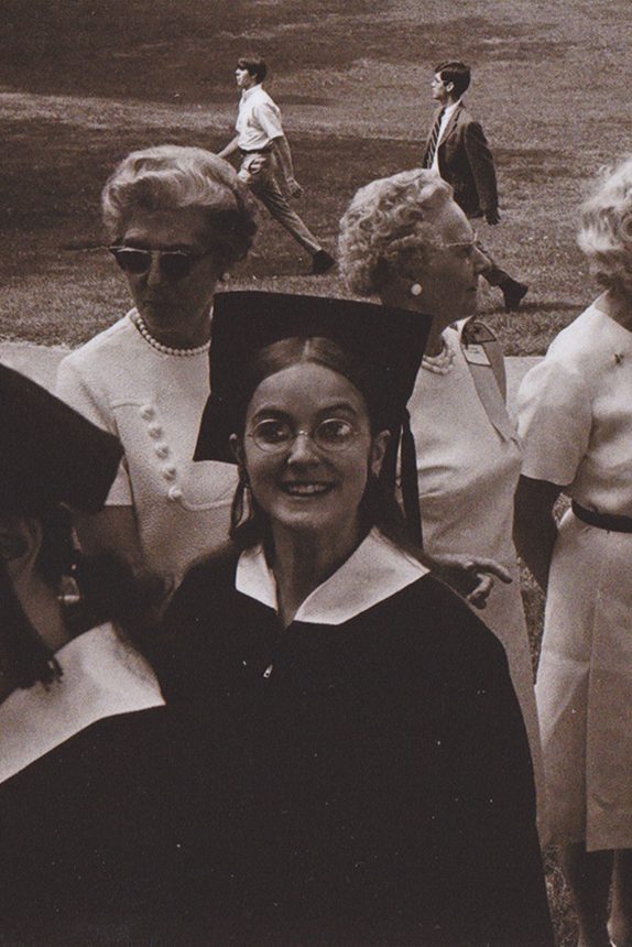 A young woman wearing her commencement gown and mortar board looks up at the camera as she processes during graduation. Behind her is a line of alumnae dressed in white.
