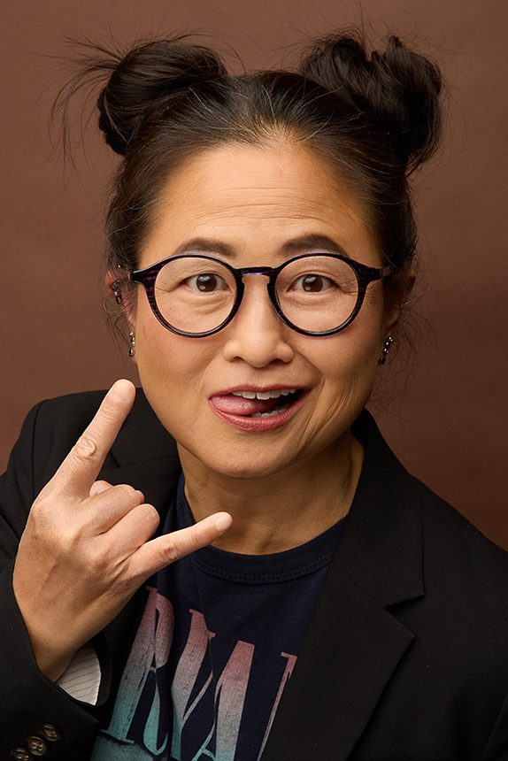 A woman wearing dark rimmed circular glasses with her hair in two buns on each side of her head holds her hand with her index and pinkie fingers extended in the horns symbol and pokes her tongue out at the camera.